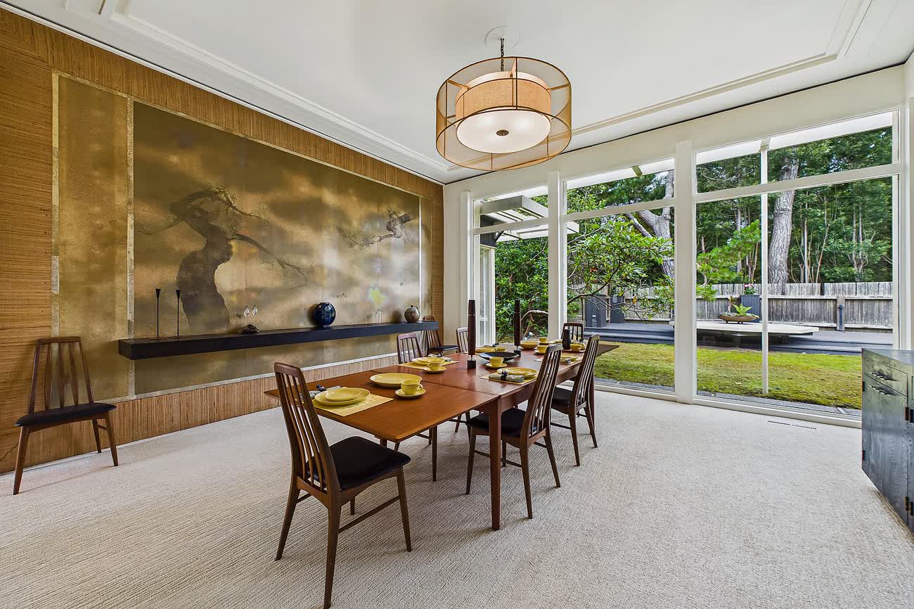 A formal dining room with a full-wall gilt mural, a long walnut table and glass on two sides opening to trees.