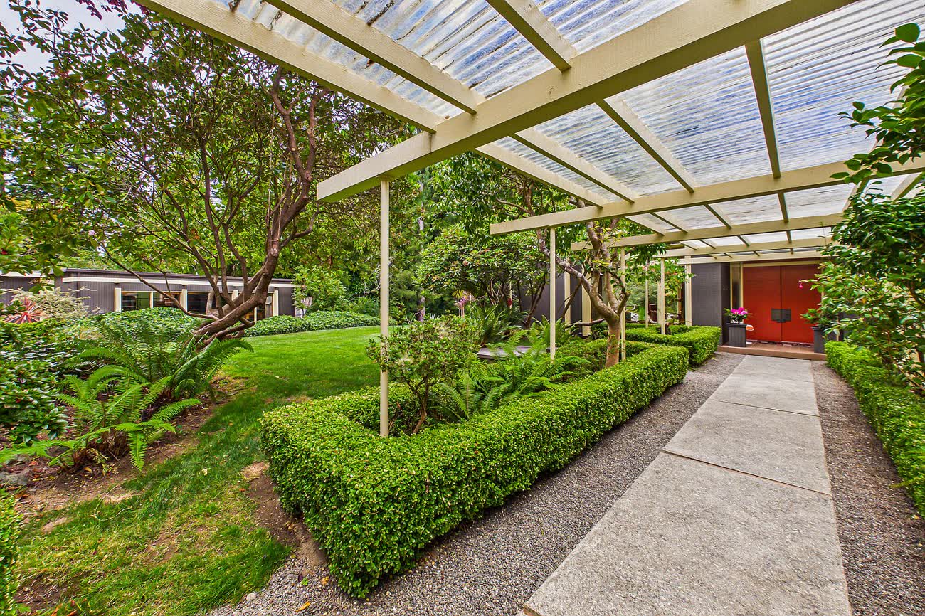 A long covered walkway with a slatted white canopy running between clipped hedges toward a red front door.